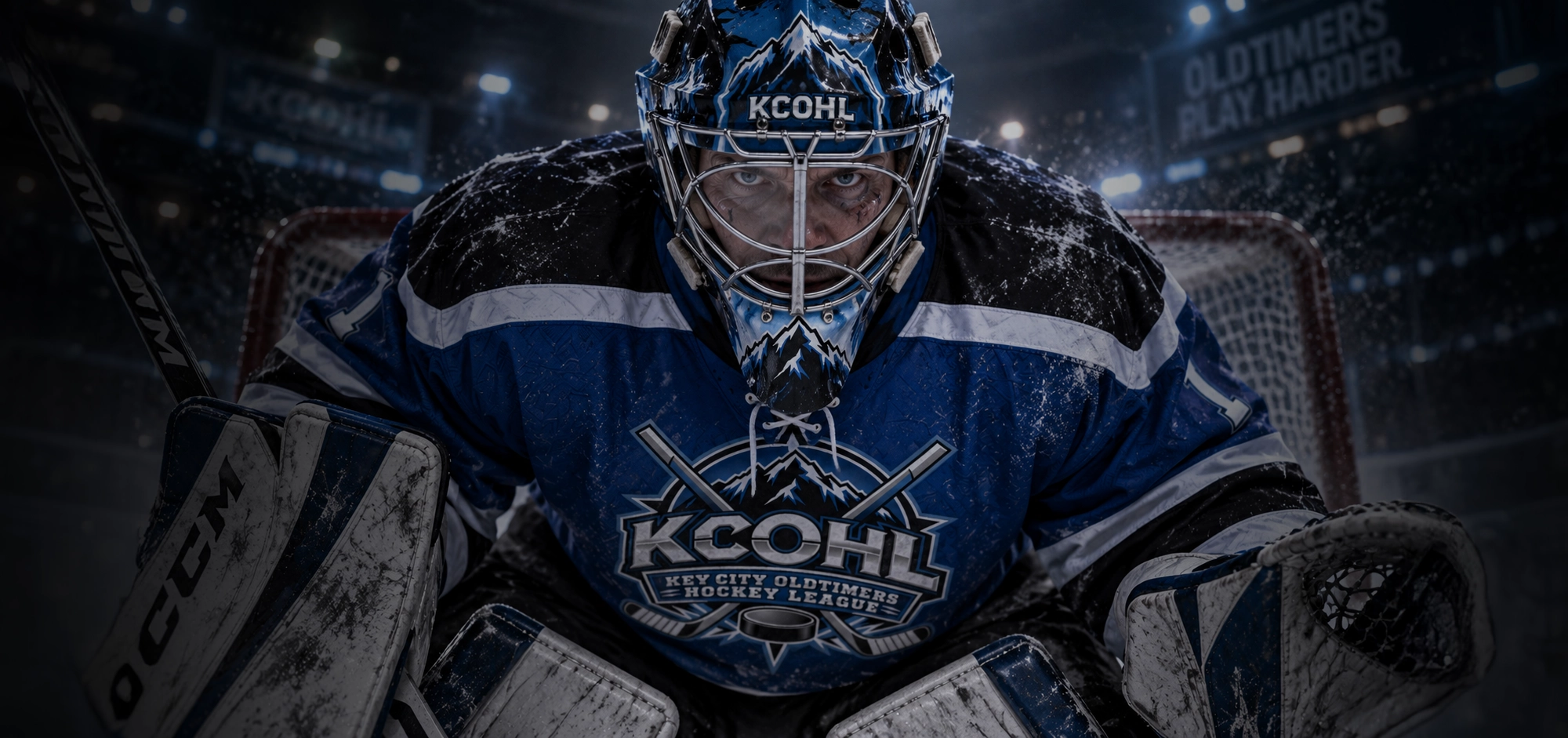 A KCOHL goalie in a blue jersey crouched in the crease, eyes on the shooter